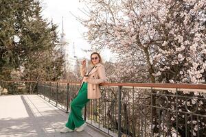 une femme est permanent sur une rebord avec une tasse de café dans sa main. elle est portant des lunettes de soleil et une bronzer manteau. le scène est ensemble dans une parc avec Cerise fleurs dans floraison. le femme est profiter le voir. photo