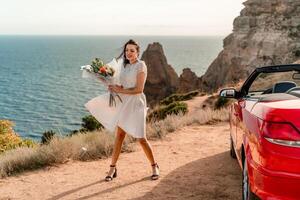 une femme dans une blanc robe est permanent suivant à une rouge convertible auto. elle est en portant une bouquet de fleurs et elle est dans une content ambiance. le scène suggère une romantique ou spécial occasion. photo