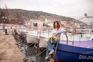 femme détient Jaune tulipes dans port avec bateaux amarré dans le arrière-plan., couvert jour, Jaune chandail, montagnes photo