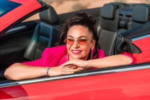 une femme dans une rose veste est séance dans une rouge convertible. elle est portant des lunettes de soleil et a sa main sur sa hanche. scène est détendu et insouciant. photo