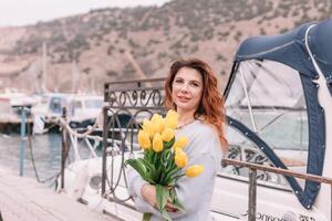 femme détient Jaune tulipes dans port avec bateaux amarré dans le arrière-plan., couvert jour, Jaune chandail, montagnes photo