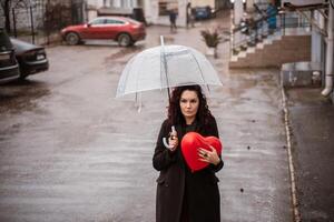 une femme en portant une rouge cœur en dessous de une clair parapluie. le parapluie est ouvert et le femme est permanent dans le pluie. photo