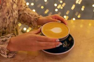 proche en haut tasse de latté café dans une café magasin. femelle mains tenir une tasse de café avec mousse latté dans le forme de une cœur sur une table dans une café. sur le arrière-plan, le lumières sont dans se brouiller. photo