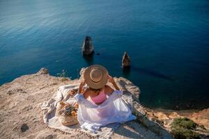 photo de une magnifique femme avec longue blond cheveux dans une rose chemise et denim short et une chapeau ayant une pique-nique sur une colline surplombant le mer