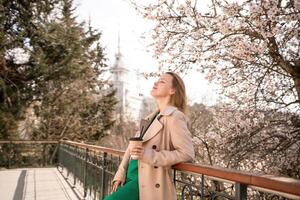 une femme est permanent sur une rebord avec une tasse de café dans sa main. elle est portant des lunettes de soleil et une bronzer manteau. le scène est ensemble dans une parc avec Cerise fleurs dans floraison. le femme est profiter le voir. photo