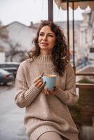 une âge moyen femme dans une beige chandail avec une bleu agresser dans sa mains est dans une rue café sur le véranda photo