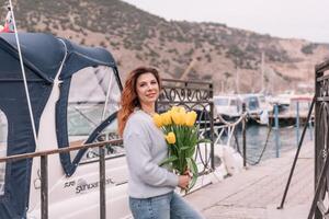 femme détient Jaune tulipes dans port avec bateaux amarré dans le arrière-plan., couvert jour, Jaune chandail, montagnes photo