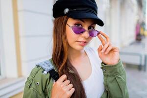 content brunette femme posant Extérieur . élégant chapeau, des lunettes de soleil et vert veste. photo