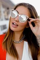 blond femme dans gros des lunettes de soleil avec plein lèvres posant Extérieur. rouge veste, élégant argent accessoires. photo