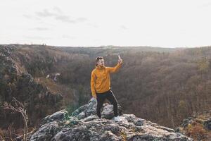 voyageur dans une Jaune veste permanent sur le bord de une falaise prise une selfie sur le sien mobile téléphone à le coucher du soleil. divoka sarka vallée, Prague photo