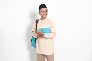 portrait de excité étudiant asiatique musulman homme dans koko chemise avec calotte porter sac à dos, tandis que en portant le sien école livres. islamique éducation concept. isolé image sur blanc Contexte photo