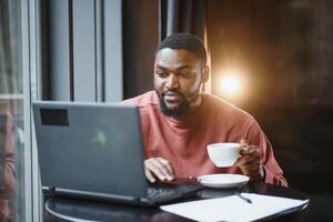 portrait de réfléchi et concentré Jeune africain américain rédacteur, travail sur Nouveau des articles en utilisant le sien ordinateur portable, et, à la recherche à papiers sur une café tableau. photo