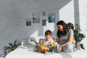 maman avec fils dans pyjamas en mangeant fruit sur lit. portrait content, souriant, joyeux famille, mère et fils ayant petit déjeuner dans lit, surprise sur maman journée. positif Humain émotions photo