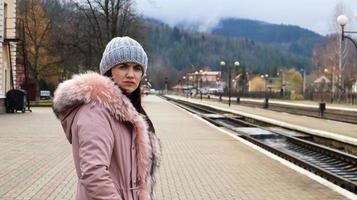 une femme vêtue d'un bonnet gris et d'une veste rose avec fourrure attend le train à la gare en regardant le train qui arrive. Transport ferroviaire. concept de voyage d'automne et d'hiver. photo