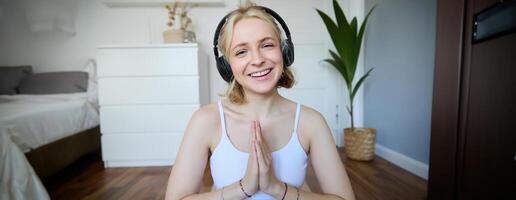 portrait de Jeune détendu femme, est assis dans pièce dans écouteurs, fermoir mains ensemble, médité, écoute à yoga la musique photo