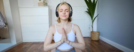 portrait de Jeune détendu femme, est assis dans pièce dans écouteurs, fermoir mains ensemble, médité, écoute à yoga la musique photo