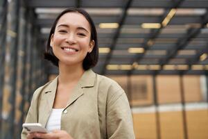 La technologie et personnes. souriant asiatique fille avec mobile téléphone, en utilisant Téléphone et en marchant dans ville dans lumière du jour photo