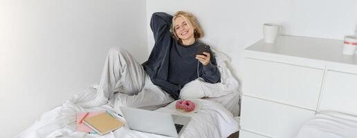 portrait de Jeune magnifique blond femme, sentiment paresseux à maison, mensonge dans lit avec tasse de thé, Donut et ordinateur portable, en train d'étudier à distance ou en train de regarder film en ligne photo