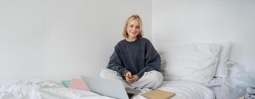 portrait de femelle étudiant, femme est assis sur lit avec portable et des cahiers, en train d'étudier en ligne, éloigné éducation concept. fille pigiste travail de sa chambre photo