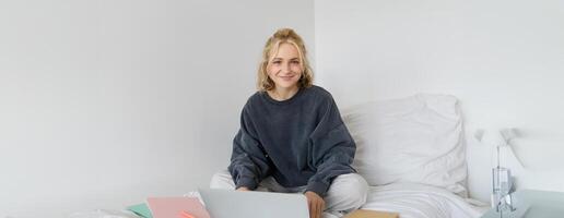 image de content Jeune femme, étudiant e-learning de maison, relier à en ligne cours sur sa ordinateur portable, est assis sur lit avec des cahiers, souriant et à la recherche content photo