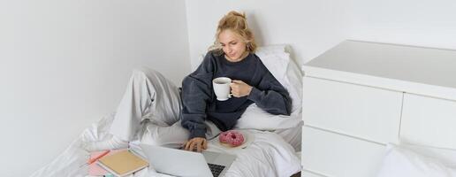 portrait de Jeune magnifique blond femme, sentiment paresseux à maison, mensonge dans lit avec tasse de thé, Donut et ordinateur portable, en train d'étudier à distance ou en train de regarder film en ligne photo