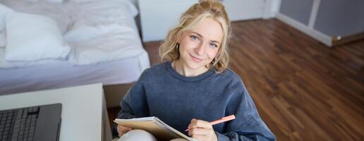 proche en haut portrait de magnifique Jeune femme, l'écriture dans sa agenda, journal ou planificateur, séance dans une chambre, souriant et à la recherche à caméra photo