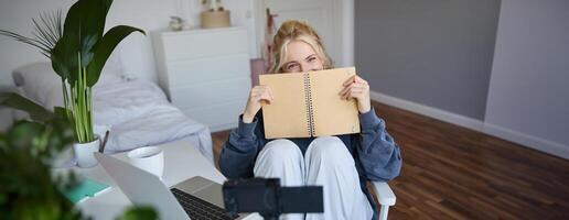 portrait de mignon, souriant Jeune social médias contenu créateur, fille enregistrements sur numérique caméra et stabilisateur, détient carnet de notes, pourparlers à public, vlogging dans sa pièce photo