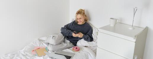 mode de vie et gens concept. Jeune magnifique femme, rester à maison, mensonge dans lit avec portable et téléphone intelligent, en mangeant Donut, profiter gratuit temps, dépenses fin de semaine à maison, en train de regarder film en ligne photo