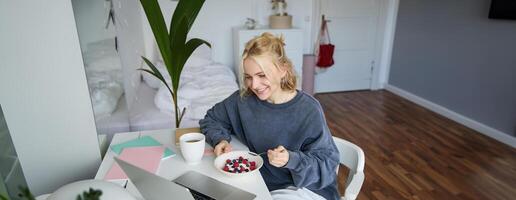 image de souriant Jeune femme en train de regarder s sur portable tandis que en mangeant petit déjeuner et en buvant thé, à la recherche à filtrer, séance dans sa pièce photo