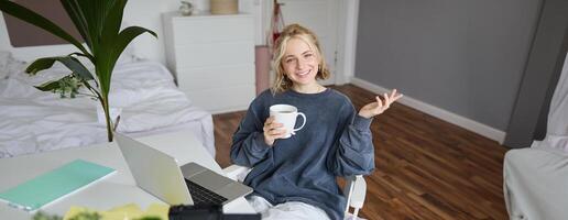 image de Jeune femme, social médias influenceur, édition sa sur ordinateur portable, est assis dans une pièce avec ordinateur et numérique caméra, en buvant café, souriant à caméra photo