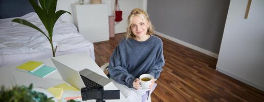 portrait de Jeune souriant femme, social médias influenceur, fille avec numérique caméra et ordinateur portable, est assis avec tasse de thé dans chambre à coucher, enregistrements vlog, crée contenu photo