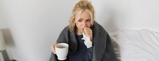 portrait de femme contagieux une froid, rester Accueil malade, en buvant thé, éternuements dans serviette de table, soufflant nez, a convoitise symptômes photo