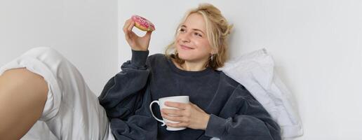 proche en haut portrait de magnifique souriant, blond femme, mensonge dans lit, les boissons thé et mange Donut sur une paresseux fin de semaine ou jour de congé photo