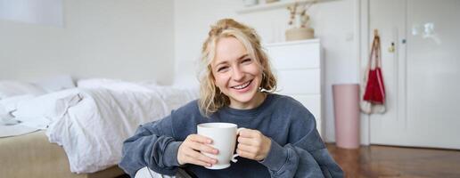 portrait de Jeune femme séance sur chambre sol, en buvant thé, en portant blanc agresser et souriant à caméra photo