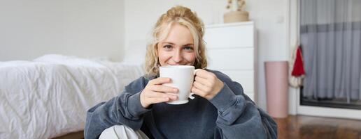 image de Jeune adolescent fille séance dans sa chambre sur sol, en buvant tasse de thé et profiter journée à maison, souriant et à la recherche à caméra photo