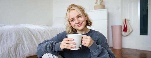 image de Jeune adolescent fille séance dans sa chambre sur sol, en buvant tasse de thé et profiter journée à maison, souriant et à la recherche à caméra photo