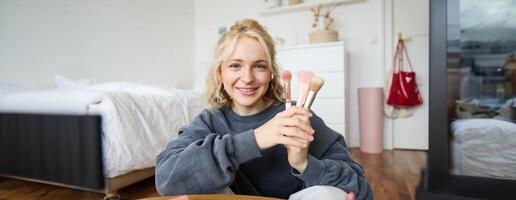 portrait de Jeune femme, contenu créateur, fabrication une à propos se maquiller, montrant brosses à public, à la recherche à caméra, enregistrement beauté Didacticiel, souriant Heureusement photo