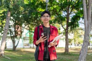 Jeune asiatique Université étudiant en utilisant téléphone intelligent avec content expression. une Masculin souriant tandis que en portant le sien téléphone et livres à le Publique parc. copie espace photo