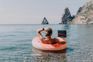 femme pigiste travaux sur portable nager dans mer sur rose gonflable anneau. content touristique flottant sur gonflable Donut et travail sur portable ordinateur dans calme océan. free-lance, éloigné travail nulle part photo