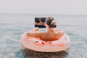 femme pigiste travaux sur portable nager dans mer sur rose gonflable anneau. content touristique flottant sur gonflable Donut et travail sur portable ordinateur dans calme océan. free-lance, éloigné travail nulle part photo