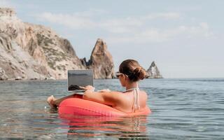 femme pigiste travaux sur portable nager dans mer sur rose gonflable anneau. content touristique dans des lunettes de soleil flottant sur gonflable Donut et travail sur portable ordinateur dans calme océan. éloigné travail nulle part photo