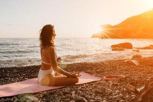 femme mer yoga. content femme dans blanc maillot de bain et boho style bracelets pratiquant en plein air sur yoga tapis par mer sur le coucher du soleil. femmes yoga aptitude routine. en bonne santé mode de vie, harmonie et méditation photo