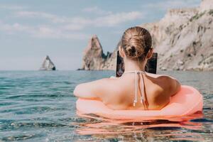 femme pigiste travaux sur portable nager dans mer sur rose gonflable anneau. content touristique dans des lunettes de soleil flottant sur gonflable Donut et travail sur portable ordinateur dans calme océan. éloigné travail nulle part photo