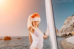 femme mer souper. proche en haut portrait de content Jeune caucasien femme avec longue cheveux dans Père Noël chapeau à la recherche à caméra et souriant. mignonne femme portrait dans une blanc bikini posant sur souper planche dans le mer photo