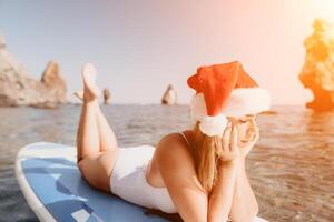 femme mer souper. proche en haut portrait de content Jeune caucasien femme avec longue cheveux dans Père Noël chapeau à la recherche à caméra et souriant. mignonne femme portrait dans une blanc bikini posant sur souper planche dans le mer photo