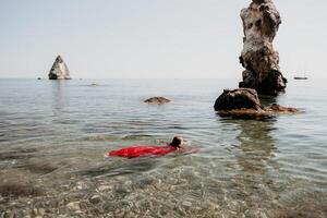 femme Voyage mer. content touristique dans rouge robe prendre plaisir prise image en plein air pour souvenirs. femme voyageur posant dans mer plage, entouré par volcanique montagnes, partage Voyage aventure périple photo