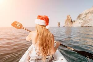 femme dans kayak retour voir. content Jeune femme dans Père Noël chapeau flottant dans kayak sur calme mer. été vacances vacances et de bonne humeur femelle gens relaxant ayant amusement sur le bateau. photo