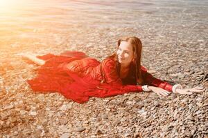 femme Voyage mer. content touristique dans rouge robe prendre plaisir prise image en plein air pour souvenirs. femme voyageur posant dans mer plage, entouré par volcanique montagnes, partage Voyage aventure périple photo