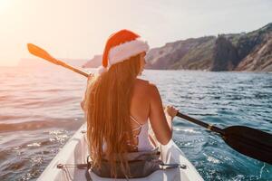 femme dans kayak retour voir. content Jeune femme dans Père Noël chapeau flottant dans kayak sur calme mer. été vacances vacances et de bonne humeur femelle gens relaxant ayant amusement sur le bateau. photo