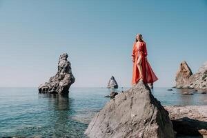 femme Voyage mer. Jeune content femme dans une longue rouge robe posant sur une plage près le mer sur Contexte de volcanique rochers, comme dans Islande, partage Voyage aventure périple photo